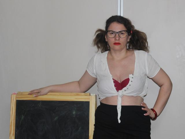 KinkyKosmina with glasses, red lipstick, and tied-up white shirt exposing red bra, black skirt, hand on hip, leaning on wooden chalkboard. Brown puffy hair, slightly overweight.