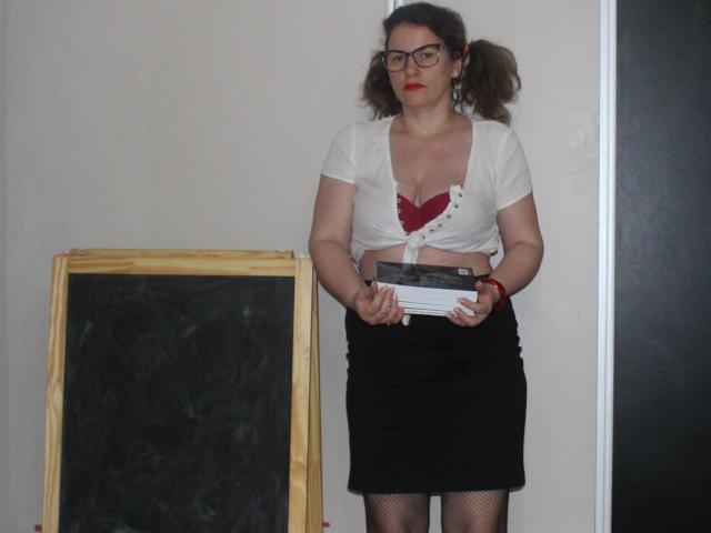 KinkyKosmina stands with puffy brown hair in pigtails, glasses, red lipstick, white tied-up shirt exposing red heart-shaped bra, black skirt, holding book, standing in front of chalkboard.