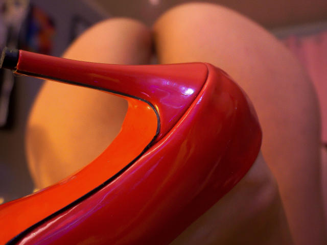 Red high heel shoe with shiny, reflective surface positioned between ZoeeMar's bare, tan-colored ass. The heel is tilted, highlighting its sleek, curved design.