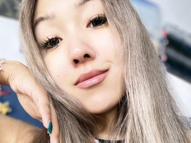 RenyLime has light brown skin, green eyes with long lashes, and shiny silver-gray hair. She's smiling with pink lipstick, green nails, and a silver bracelet on her right hand.
