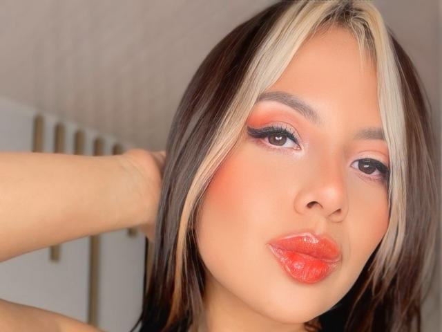 NinaDeville with blonde and brown hair, red eyeliner, and shiny red lips, pouting slightly. Her arm is raised, showing off her makeup and glossy lips.
