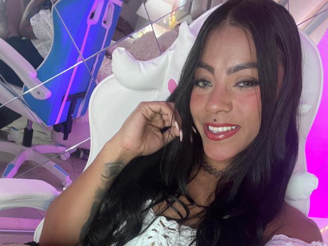 Amberv has long black hair, a nose ring, and is smiling with red lipstick. She's touching her hair with a white dress and a tattoo on her arm.