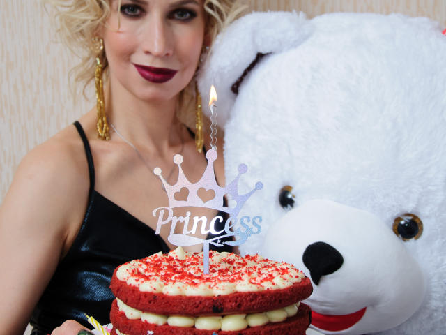 AdrianaSea with curly blonde hair, red lipstick, and gold earrings, stands next to a giant white teddy bear, holding a red cake with white frosting and a candle.