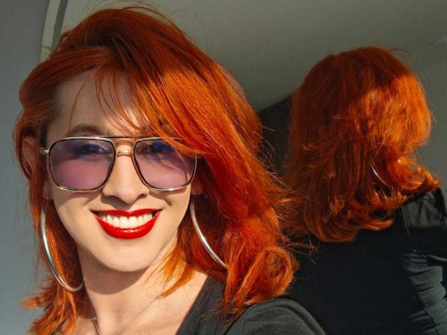JulianaSaenz with bright red hair, wearing pink sunglasses, red lipstick, and large hoop earrings, smiling at a mirror reflection showing her back. Her tits are small.