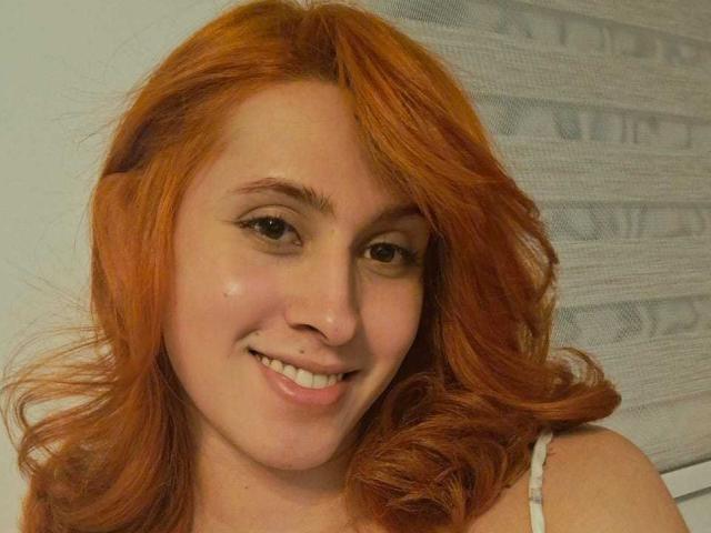JulianaSaenz has red, wavy hair, light skin, and is smiling with her small tits visible in a white top. She has a cute face and black eyeliner.