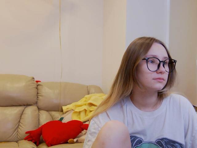 CaramellSex, a white chick with glasses and blonde hair, wearing a grey tee, sits on a beige couch with a yellow blanket and red stuffed heart.