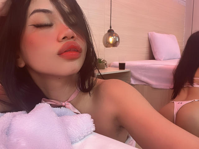 LiliMays with pink lipstick, red blush, black hair, pink halter top, holding pink and white towel, mirror reflecting her back, pink thong, soft lighting, pouty expression.
