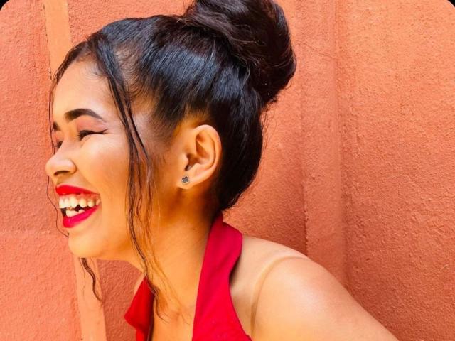 MaryamGomez with her black hair in a bun, smiling with bright red lipstick, wearing a red halter top, standing against a textured orange wall. Her ears have small studs.