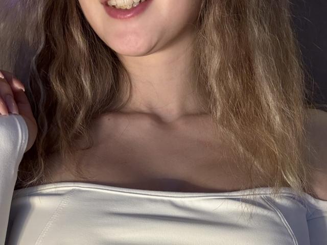 SelinaRose's upper body, showing her wavy blonde hair, white off-shoulder top, and slightly open mouth with visible teeth. Her small, pale tits are partially visible.