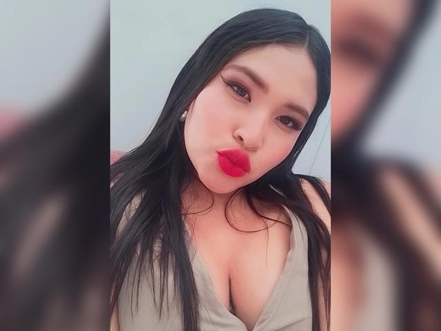 OliviGrace with long black hair, red lipstick, and a grey tank top showing her big tits, pouting her lips, and looking into the camera. Blurred reflections in the foreground.