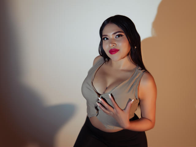 OliviGrace, con lunghi capelli neri, rossetto rosso, top senza maniche color pelle, pantaloni neri, che tiene il seno con una mano, ombra su un muro bianco, che guarda la fotocamera..