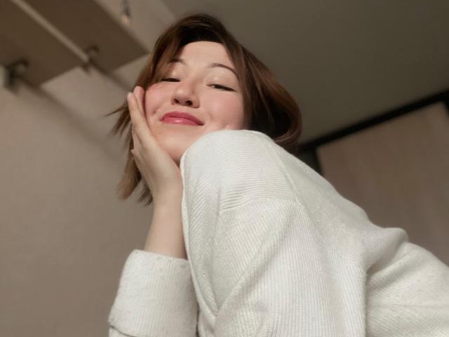 Sillitls with cute face, short brown hair, and pink lips, wearing a white sweater, smiling with hand on cheek, looking down. Sillitls' tits are not visible.