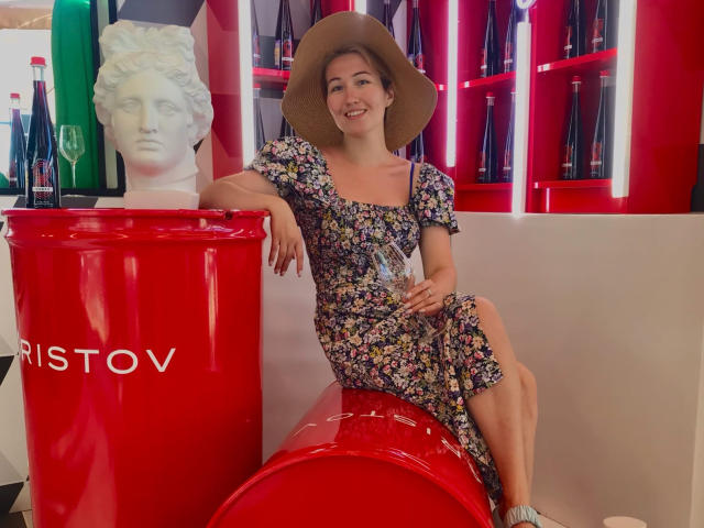Sillitls with floral dress, brown hat, and smile, sitting on red cylinder, leaning on white bottle, holding glass, next to white bust and red shelf with bottles.