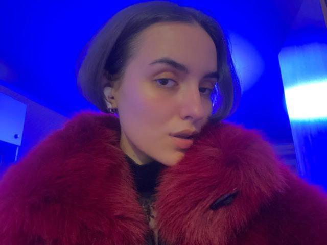 BellaSpace with short brown hair, red fur coat, and subtle makeup, looking slightly to the side, in a blue-lit environment. She's wearing earrings and has a slight smirk.