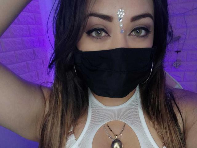 ZahraJalid with green eyes, wearing a black mask, white mesh top, and a forehead jewel. Her tits are visible through the top, with a necklace in the center.