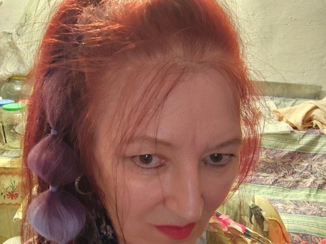 Sabrhina has red and purple hair in a loose braid, wearing red lipstick, and looking at the camera with a slight smirk. She has fair skin and small, brown eyes.