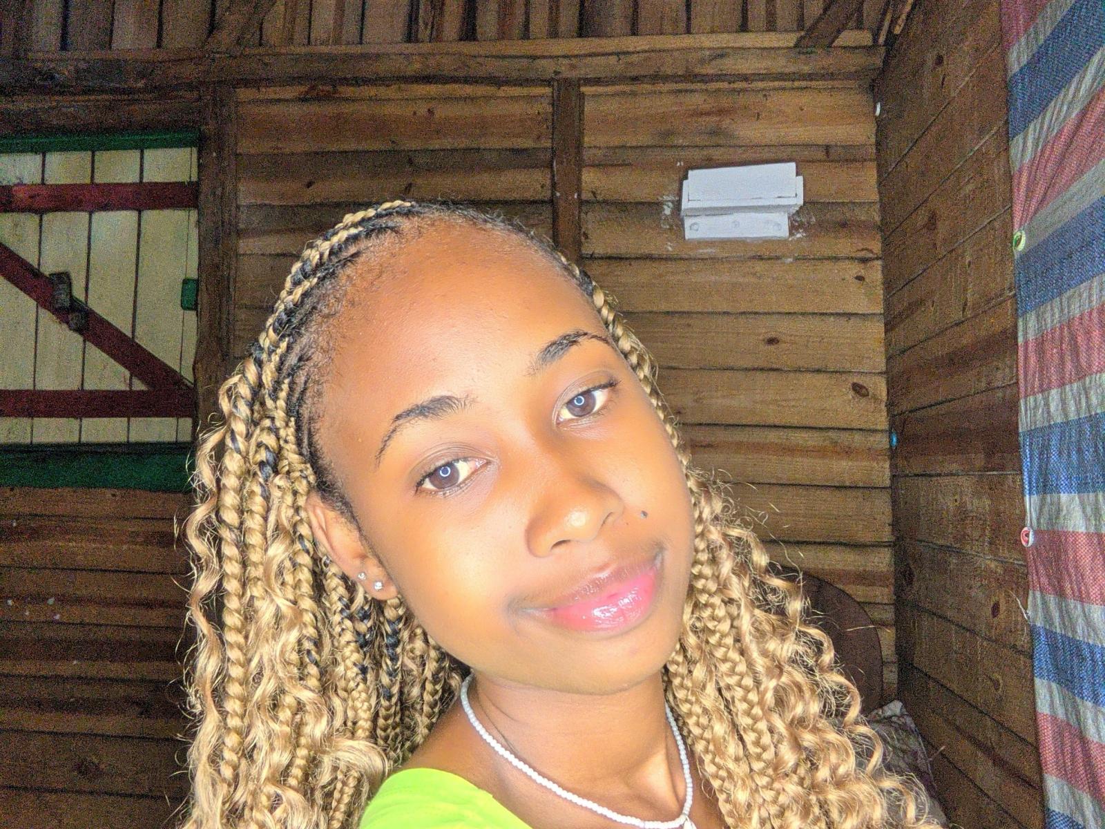ClaraNice69 with light brown skin, blonde braided hair, green shirt, and pink glossy lips, smiling softly, wearing a white necklace, in a wooden room.