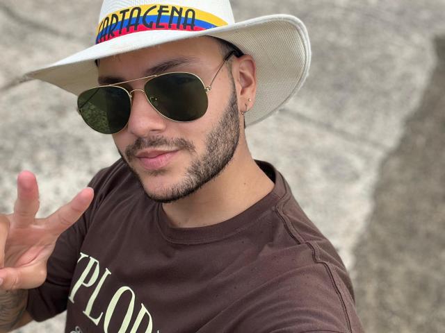 HenryEdward with a white cowboy hat, gold aviator sunglasses, brown shirt, and tattooed hand giving a peace sign, sporting a beard and small nose ring, smiling.