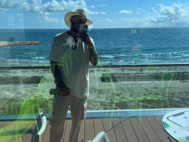 HenryEdward, standing in front of a glass window, wearing a white shirt, beige pants, and a straw hat, taking a selfie with a phone, reflecting ocean and sky.