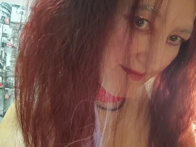 Sabrhina with messy red-brown hair, slightly parted lips, and a black choker, looking up with subtle makeup and a cute expression. Her eyes are shiny and she's blushing.