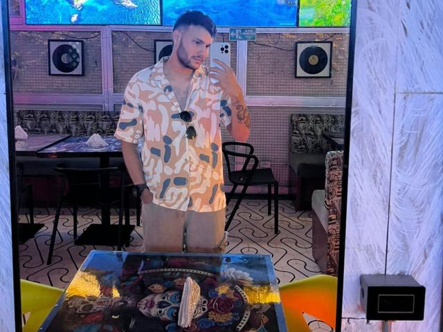 HenryEdward with a beard, wearing a patterned shirt, holding a phone, showing a tattoo on his right arm, standing in front of a mirror, with his dick not visible.