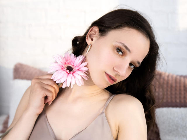 StacieLove con una flor bohemia rosa en el cabello castaño oscuro, sosteniéndola cerca de su rostro, vistiendo una camiseta sin mangas color beige, ojos marrones y aretes dorados, mirando ligeramente hacia un lado..