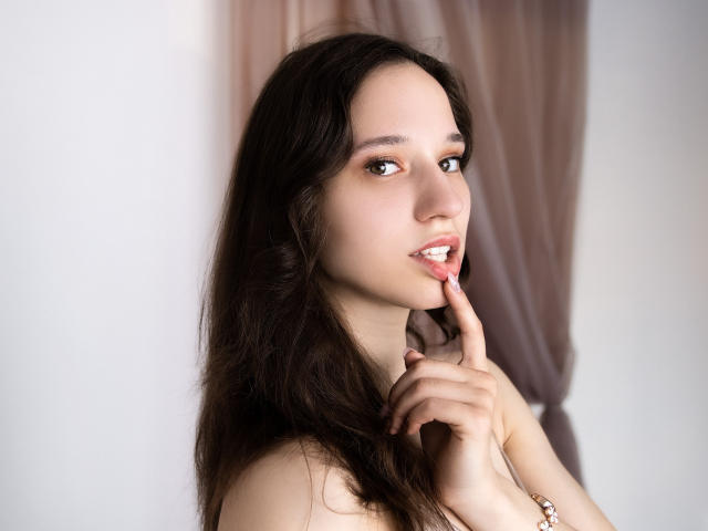 StacieLove with long brown hair, fair skin, and pink lipstick, touching her finger to her lips, looking seductive, wearing a pink bracelet, in a soft, natural light.