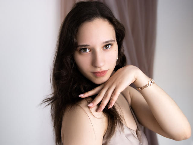 StacieLove, com cabelo castanho, olhos castanhos e pele clara, vestindo um vestido bege, tem a mão perto do pescoço, mostrando uma pulseira de ouro, e um sorriso sutil e sexy..