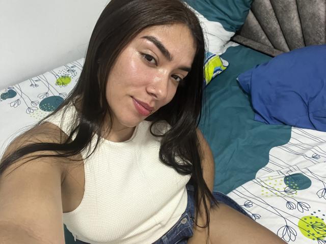 SharonKlart with long black hair, wearing a white ribbed tank top and blue shorts, sitting on a bed with a slight smile, showing her cute face and small nose.