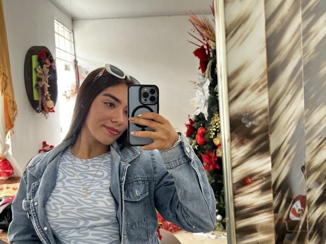 SharonKlart with long dark hair, blue-eyed, wearing a denim jacket over a white and blue patterned top, holding a phone, smiling, in a room with Christmas decorations.
