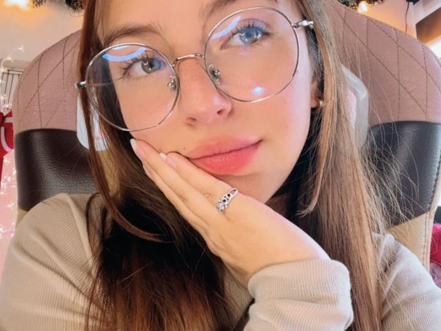MillyKenner con gafas, mano en la mejilla, labios rosados, cabello castaño, vistiendo un suéter gris, anillo en el dedo, luciendo linda, expresión suave, reflejo en las gafas..