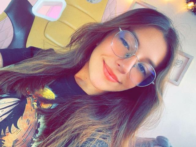 MillyKenner tiene el pelo largo y castaño, usa gafas redondas y una camisa negra con gráficos coloridos, sonríe con lápiz labial rojo, mostrando su cara linda y su pequeña nariz..