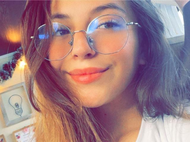 MillyKenner tem óculos redondos e charmosos, cabelo castanho claro ondulado, e está sorrindo com batom vermelho. Seu nariz é pequeno e sua pele é bronzeada. Ela está vestindo uma camisa branca..