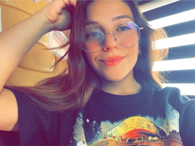 MillyKenner with long brown hair, wearing round glasses and a black tie-dye shirt, smiling with red lipstick, hand in hair, sunlight from right.
