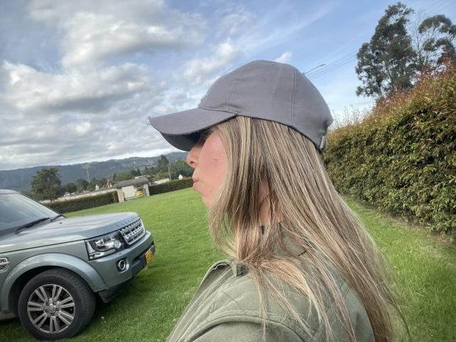 JennyLawry with blonde hair, green jacket, grey cap, side profile, looking at grey SUV on grass, cloudy sky, green hedge, trees in background.