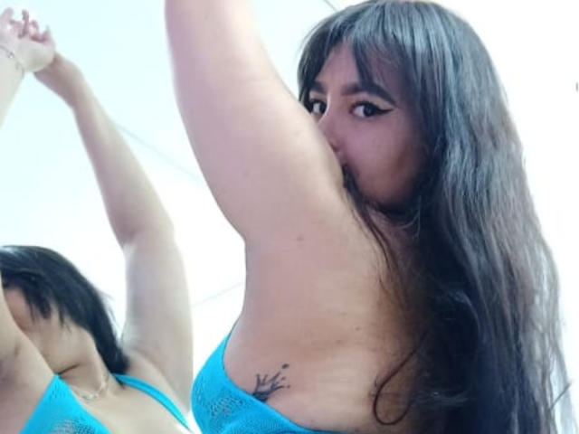 BrianaRogers with long brown hair, blue lace bra, arm raised, showing armpit tattoo, and her reflection in a mirror. Her tits are partially visible.