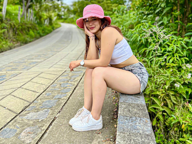LeylakWith, with pink bucket hat, white crop top, and gray shorts, sits on a stone path, legs crossed, showing her ass, wearing white sneakers, smiling.