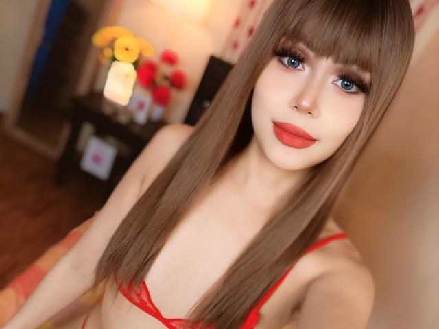 TSAnabelleSwift with long brown hair, blue eyes, and red lipstick, wearing a red bra, has a slim build and small tits, sitting in a warmly lit room.