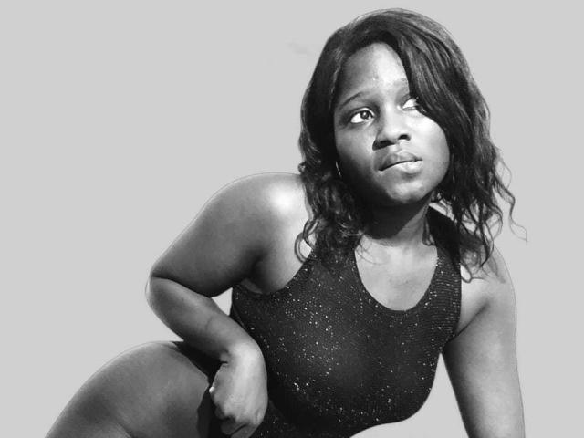 RubyMoon, a curvy black woman with medium-sized tits, wearing a sparkly black one-piece, crouches down, looking to the side with a slightly worried expression, in a black-and-white photo.