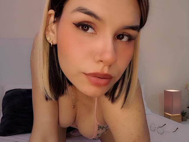 LenaBranch met blond en zwart haar, eyeliner, een neusring en een roze beha, leunt voorover, toont haar borsten en heeft een lichte glimlach. Zachte verlichting..