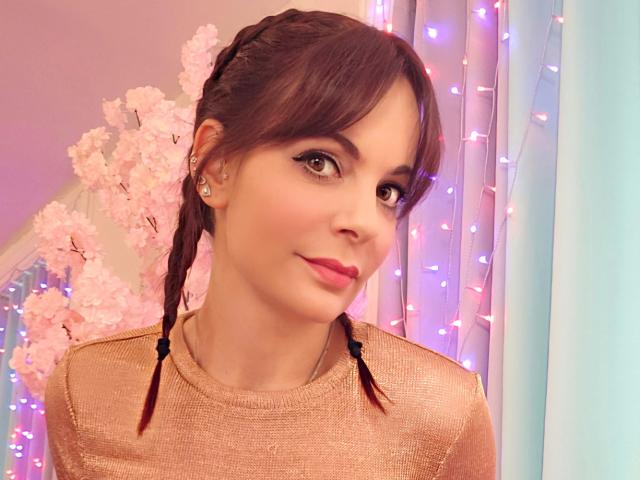 Close-up photo of JuneLaFrenchie with brown braids, wearing a shiny gold top, pink lips, and subtle makeup. Pink fairy lights and white blossoms in the background, creating a soft, romantic vibe.