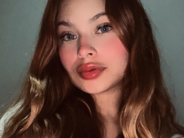 ShantalSummer has pouty red lips, blue eyes, and wavy brown hair with blonde highlights. Her face is flushed pink, and she's looking straight at the camera.