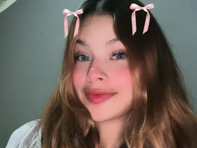 ShantalSummer has blue eyes, pink ribbons in her brown hair, and a cute smile with red lipstick. She's looking at the camera with a slight blush on her cheeks.