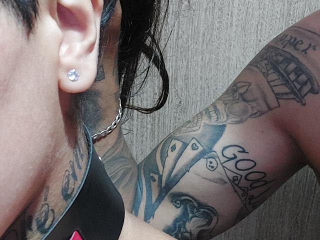 SenhorESenhoraSantos' arm tattooed with cards and text, wearing a black top, chain necklace, and diamond ear piercing, with visible ear and upper chest.