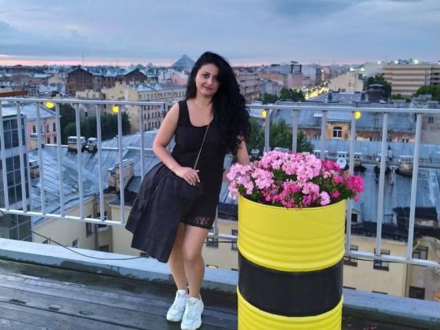 Elinor Lindy, con cabello negro largo, vestida con un vestido negro, de pie en la azotea, sosteniendo una maceta amarilla y negra con flores rosas, y zapatillas blancas..