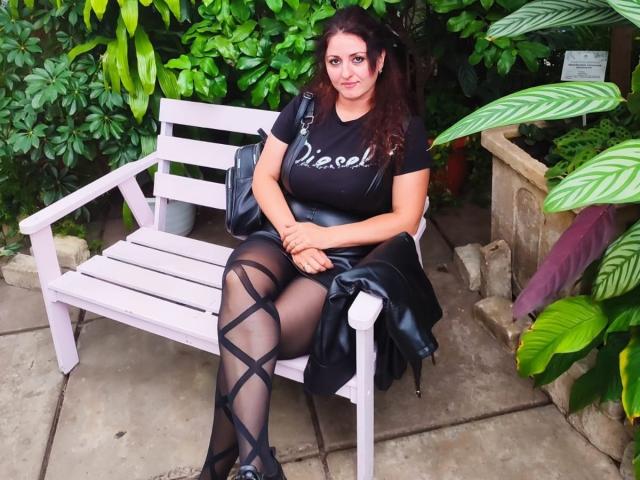 ElinorLindy with dark red hair, black "Viacel" top, black leather jacket, black criss-cross pantyhose, and black shoes, sitting on a white bench, green foliage in background.
