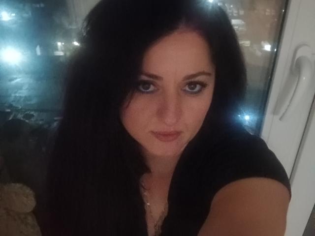 ElinorLindy has black hair, blue eyes, and a subtle smile. She's wearing a black top, and her small tits are slightly visible. She's looking directly at the camera.