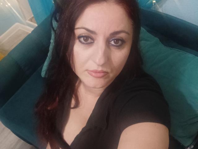 ElinorLindy tiene el cabello de color rojo oscuro, delineador de ojos negro, y lleva una blusa negra, mostrando ligeramente sus senos en una selfie. Está mirando a la cámara con una expresión neutra..