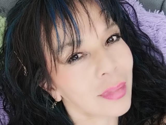 SamaraBellet's face with black wavy hair, blue highlights, brown eyes, pink lipstick, and small earrings, looking at the camera with a slight smile. She's a cute bitch.