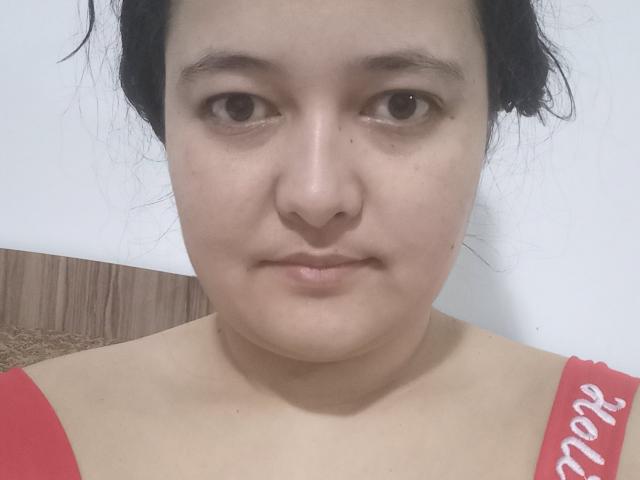 MollyJanette's face, showing a neutral expression with brown eyes, light skin, and black hair in a messy updo. She's wearing a red top with white text.